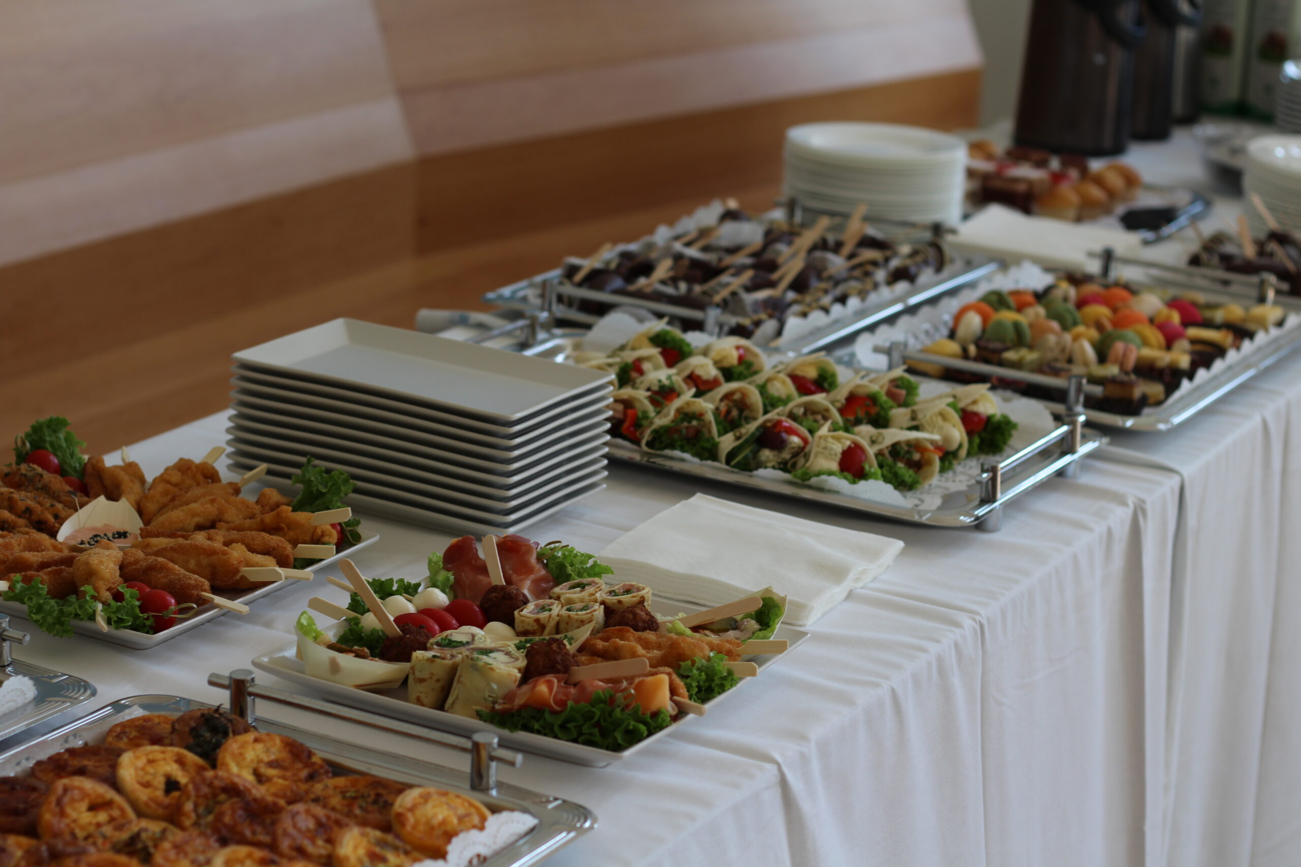 Business-Buffet mit Fingerfood-Spießen, Wraps, Desserts und gestapelten Tellern auf weißer Tafel.