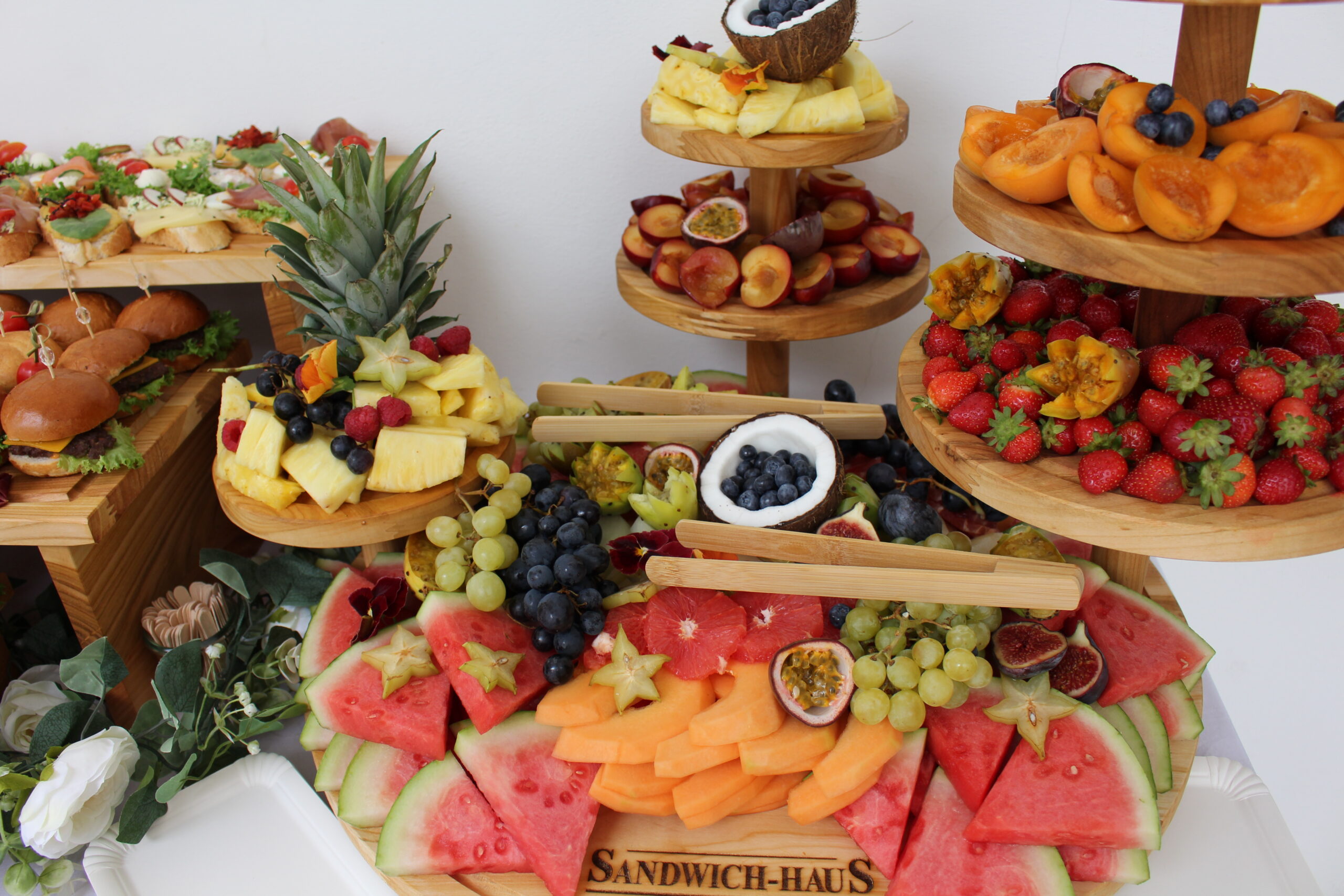 Farbige Obststation auf Holz­etageren mit Melone, Ananas, Beeren, Marillen, Trauben und Passionsfrucht – festlich dekoriert.