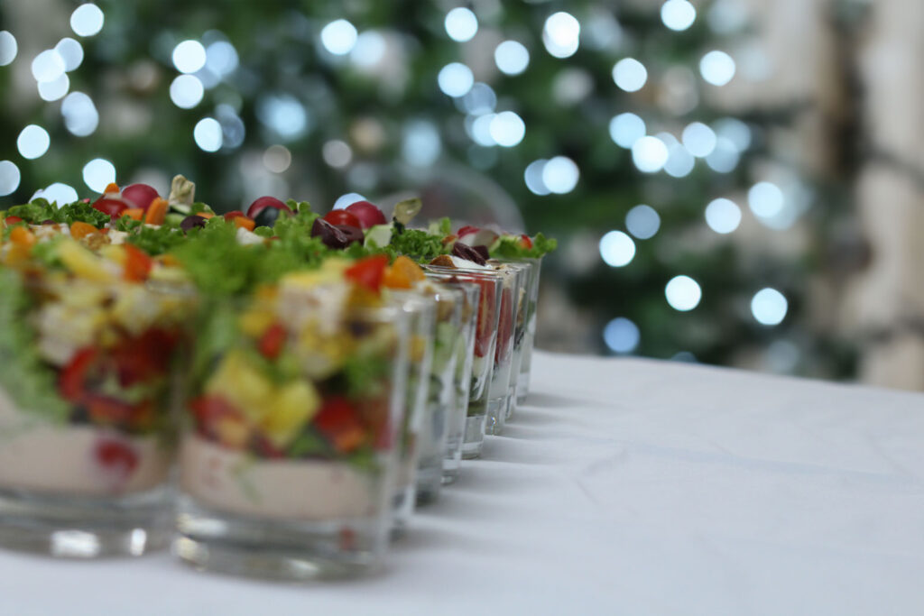 Reihe kleiner Vorspeisengläser mit Salat und Gemüse, Bokeh-Lichter eines Weihnachtsbaums im Hintergrund.