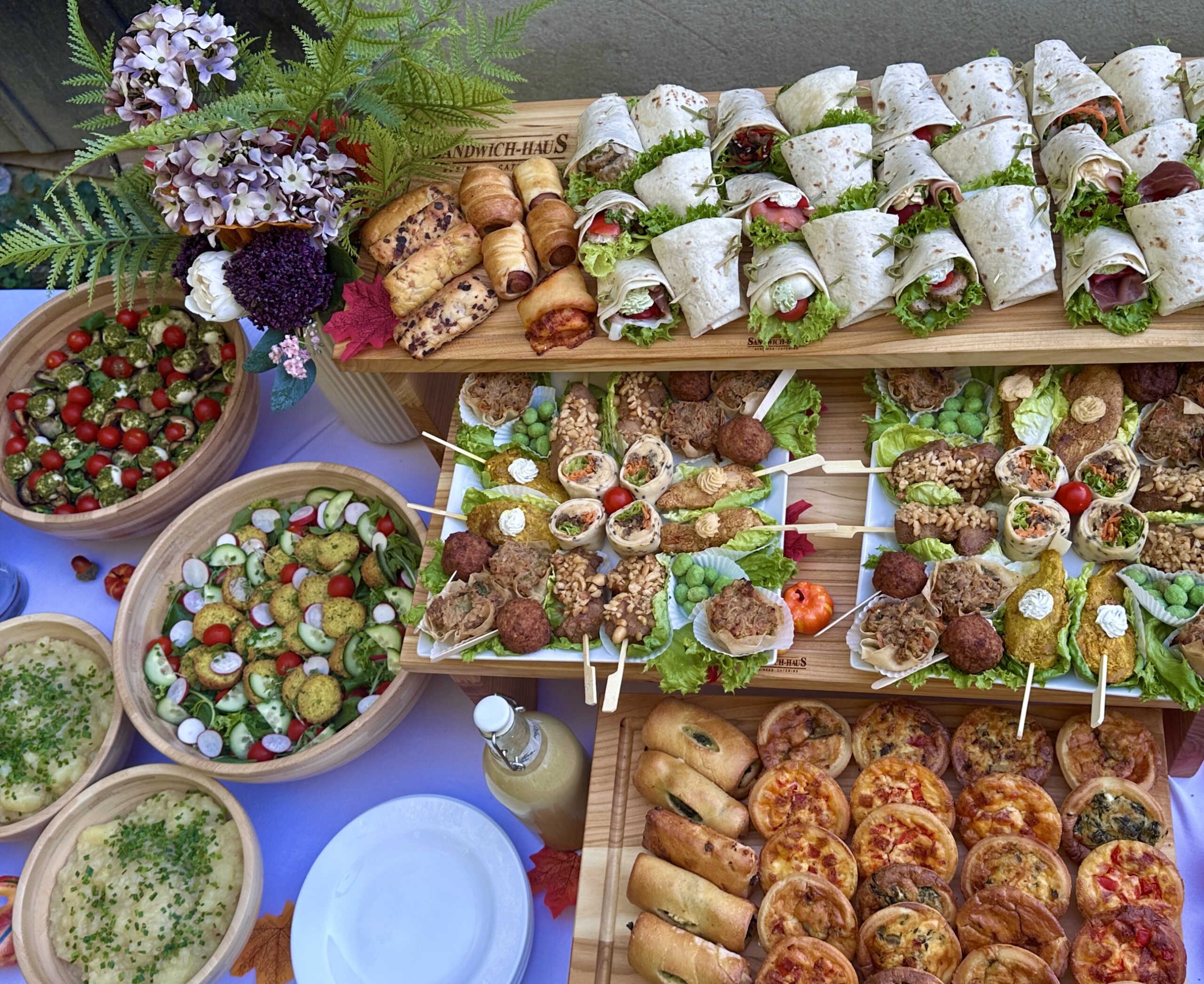 Reich gedecktes Sommerfest-Catering mit Wraps, Salatschalen, Fingerfood-Spießen, Mini-Quiches und pikantem Gebäck auf Holzetageren.